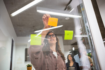 Creative woman attaching sticky notes on a glass wall during a brainstorming session with her team in a modern coworking office