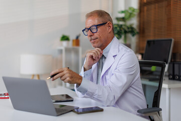 Senior middle aged male medical worker in modern clinic wearing white doctor's coat using laptop writing health personal data, consulting remotely. Telemedicine healthcare concept.