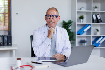 Senior middle aged male medical worker in modern clinic wearing white doctor's coat using laptop writing health personal data, consulting remotely. Telemedicine healthcare concept.
