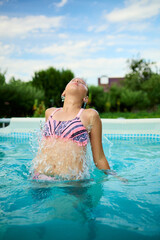 Teen girl enjoying a summer day splashing water while swimming in a pool surrounded by greenery under a bright blue sky