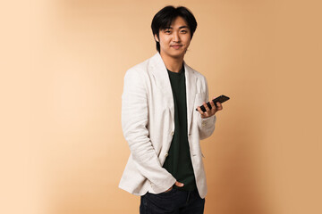 Portrait of handsome Asian businessman in suit with smartphone on beige background