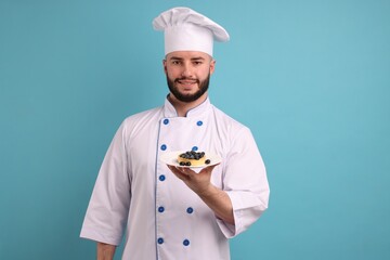 Happy confectioner in uniform holding tart of blueberries on light blue background