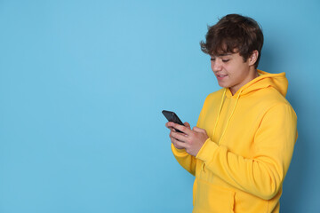 Smiling teenage boy using smartphone on light blue background. Space for text