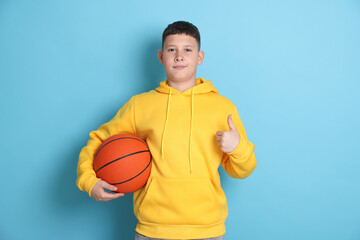 Teenage boy with basketball ball showing thumbs up on light blue background