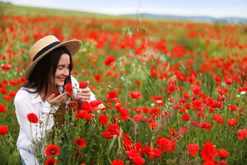Smiling woman in hat sniffing red poppy flowers in wildflower meadow