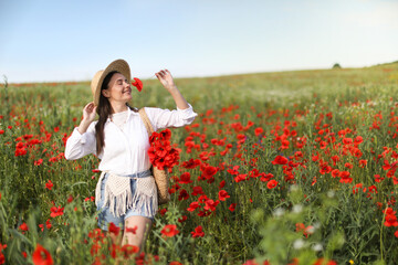 Smiling woman in hat with bag sniffing poppy flower in morning wildflower meadow