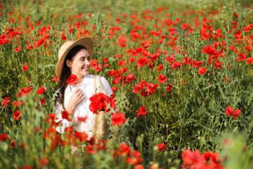 Smiling woman in hat with bag among red poppy flowers in morning wildflower meadow