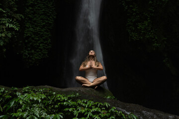 Young woman meditating in front of majestic waterfall in lush jungle