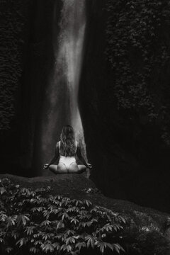 Young woman meditating in front of majestic waterfall in lush jungle