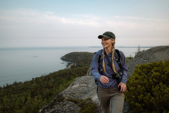 Smiling young woman hikes with camera along trail with view, Acadia