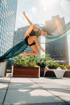 Dancer leaps as sheer fabric flows upward on city rooftop.