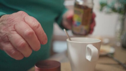 Close-up of elderly woman stirring coffee or tea with spoon, adding sugar from jar. Cozy kitchen setting with homemade breakfast, focus on daily routine and homemaking activities in morning light