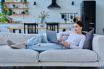 Portrait of young woman lying on sofa typing on laptop, in home interior