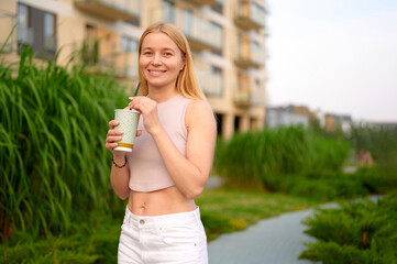smiling woman enjoying beverage outdoors on sunny day, surrounded by lush greenery and modern buildings in urban environment. casual lifestyle, summertime relaxation, carefree moment