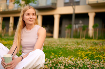 woman relaxing in green park, holding a drink and enjoying nature. casual summer outfit suggests leisure and tranquility in urban setting. blossoming plants create natural calm.