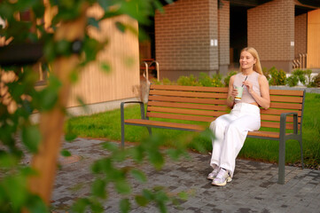 woman enjoying iced drink on outdoor bench in sunny park. casual relaxation and tranquil setting highlight leisure moments in urban nature. perfect for depicting lifestyle and outdoor vibes