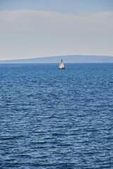 Fototapeta premium White sailboat sails calmly on open blue sea with gentle ripples, distant hills under a soft sky add depth and serenity; sailboat, seascape;