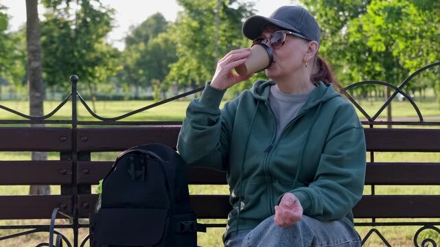 Woman in baseball cap and sportswear sits and drinks coffee in park on bench. Video 4k