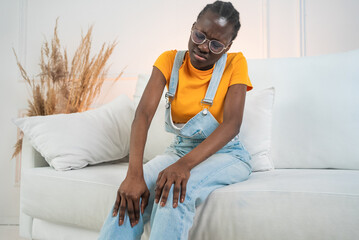 Young woman wearing casual clothes and eyeglasses is sitting on a white sofa, grimacing in pain as...