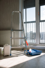 Metal stepladder stands next to paint roller and tray, buckets, and radiator in room with window...