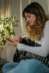 Young female musician tuning her acoustic guitar using a mobile phone app, sitting comfortably at...