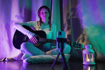 Young woman musician is sitting on floor and playing acoustic guitar at home, recording music video...