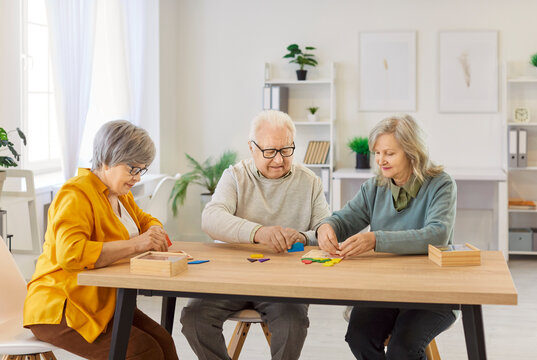 Group of elderly people sitting at wooden table, solving colorful puzzles together in nursing home. Seniors focusing on assembling pieces, engaging in fun cognitive activity for memory stimulation.