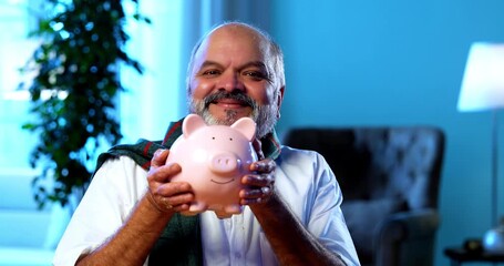 Indian senior man protecting piggy bank from imaginary thief while sitting at table in modern home, humorous concept of elderly financial security, happy aged asian man defending savings indoors