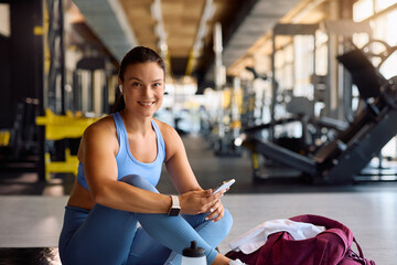 Happy athletic woman using cell phone in gym and looking at camera.