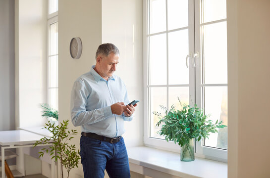 Focused senior mature man at home, looking at phone screen, standing browsing mobile device apps, reading news, shopping online, searching information in internet, holding smartphone, waiting 