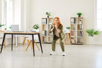 Full length portrait of young smiling woman doing stretching sit-ups exercises standing at workplace in office. Cheerful female company employee exercising for health during a break from a work.