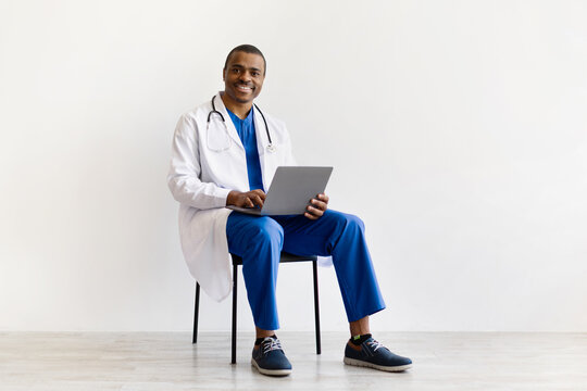 Smiling African American doctor sitting with laptop, looking at camera. Confident male healthcare professional using computer for medical tasks or telehealth work, copy space