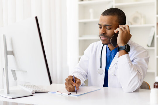 Focused African American doctor taking notes during phone conversation at work desk. Modern professional multitasking with smartphone and clipboard in bright office, closeup