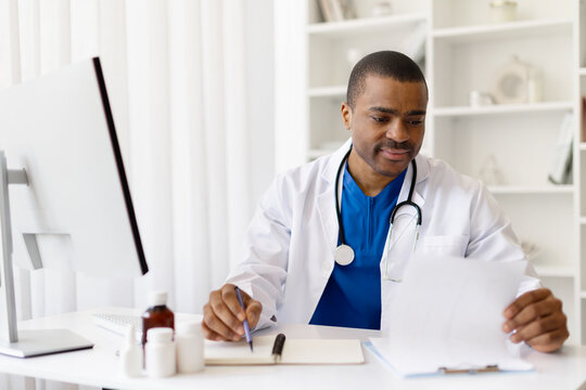 Smiling young African American doctor reading document at desk in office. Confident black male physician reviewing medical papers during workday, wearing lab coat and stethoscope