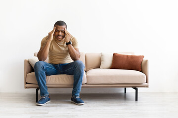 Young black man sitting on sofa with hands on temples, suffering from stress or headache. Casual...