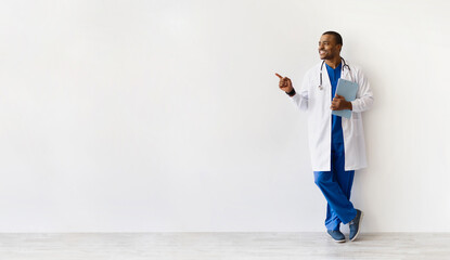 African American doctor leaning relaxed against wall, smiling and pointing aside. Confident medical worker with clipboard sharing information or recommendation, panorama