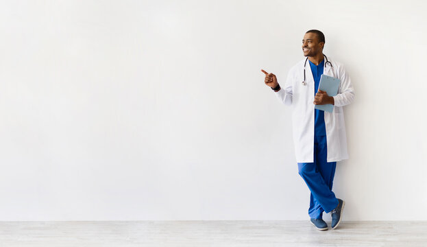 African American doctor leaning relaxed against wall, smiling and pointing aside. Confident medical worker with clipboard sharing information or recommendation, panorama