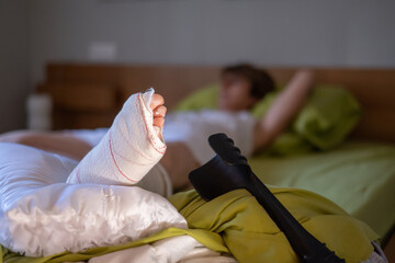 Patient is recovering in bed with a broken leg in a cast, their crutches resting nearby,...