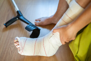 Close-up view of a bandaged foot resting on a wooden floor, accompanied by a crutch, illustrating...