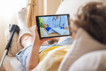 Patient with a broken leg and cast is using a tablet for a telemedicine appointment with a doctor,...