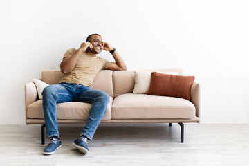 Cheerful young black man sitting on sofa, talking on smartphone and smiling. Casual and relaxed moment in home office or leisure setting with modern communication, copy space