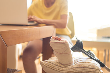 Woman with plastered leg is working on laptop while sitting at home with crutches resting on a...