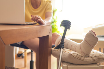 Woman managing work tasks at home, balancing a laptop on her lap while resting a plastered broken...