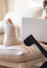 Woman with a broken leg using crutches and resting foot on a cushion, working with her laptop from...