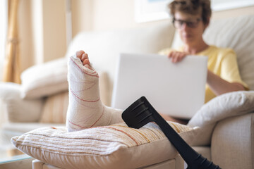 Woman with a broken leg, resting on a cushion with crutches nearby, uses her laptop, highlighting...
