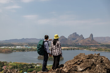 rear view of a traveling couple holding hands on a rocky summit, gazing at the landscape below that...