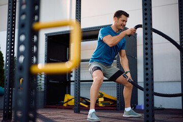 Happy athletic man working out with battle ropes in outdoor gym.