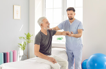 Obraz premium Male professional chiropractor or physical therapist working with mature senior man's arm sitting on the couch in rehabilitation hospital. Physiotherapy and osteopathic medicine concept.