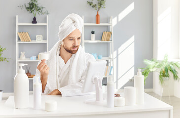 Man in a bathrobe and with a towel on head is applying face cream and looking in the mirror after a...