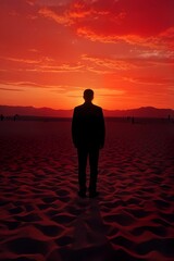 A photograph of a man turning his back, silhouetted against a vivid red and orange desert sunset. Textures of shifting sands create a bumpy foreground, while shadowy figures of distant mountains loom.